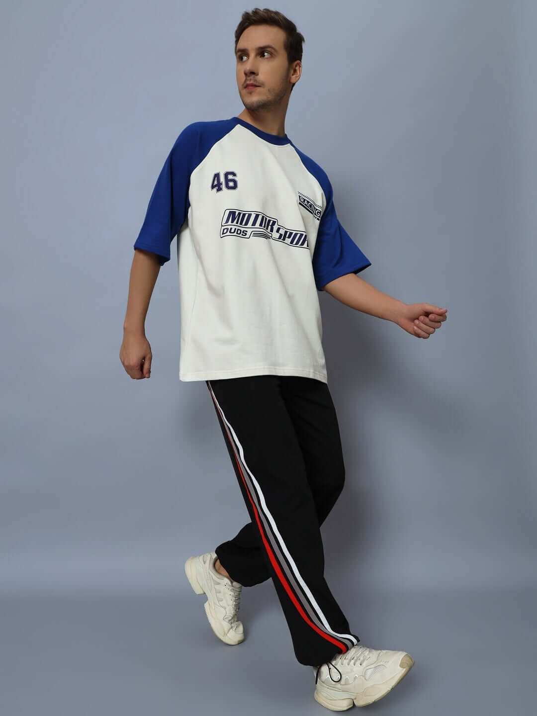 Man wearing white and blue sports shirt with black relaxed fit joggers featuring red and white side stripes against a plain background.