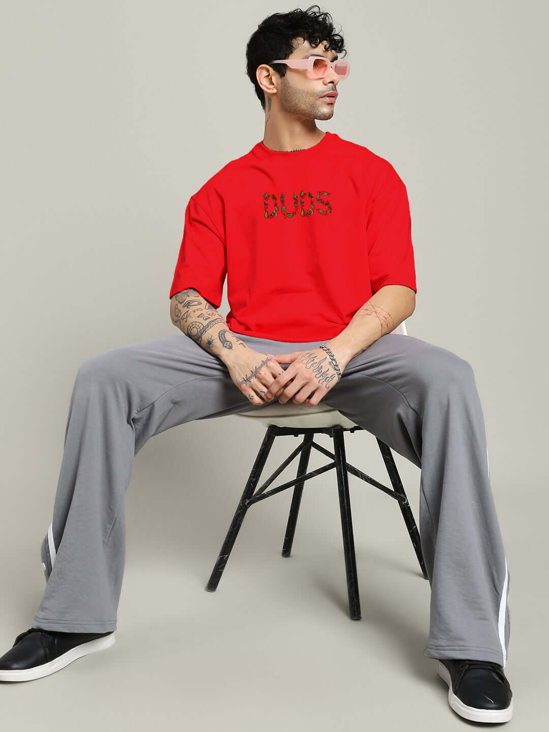 Model wearing red Dragonfly oversized T-shirt, paired with gray pants and sunglasses, sitting on a stool.