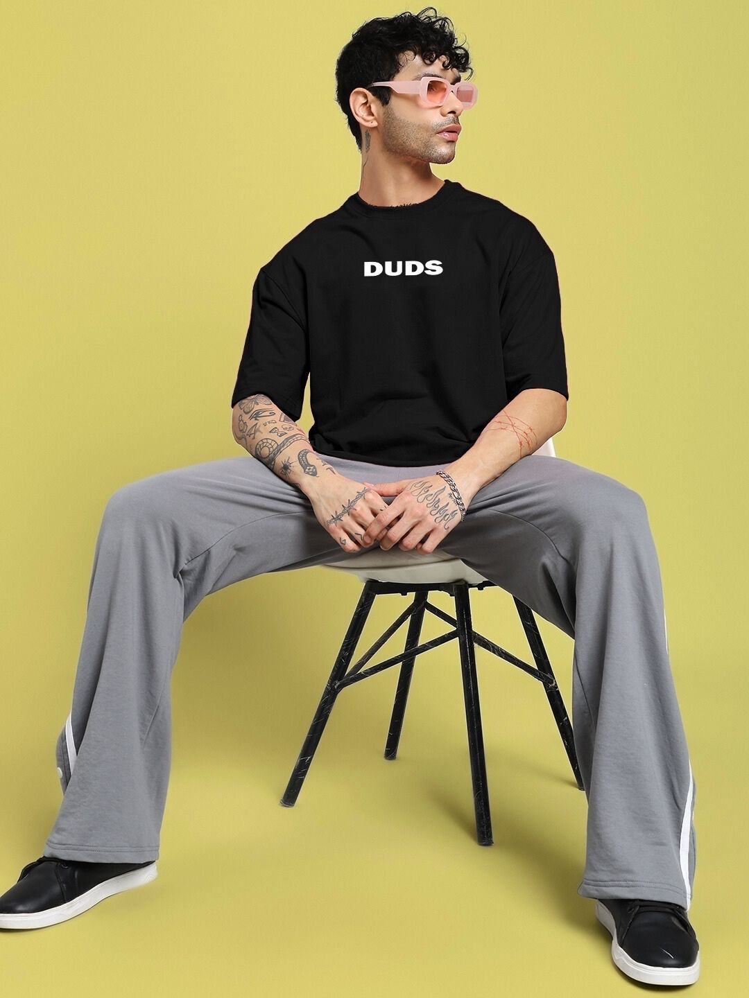 Man wearing DREAMLESS OVER-SIZED T-SHIRT BLACK sitting on chair, yellow background, stylish streetwear fashion