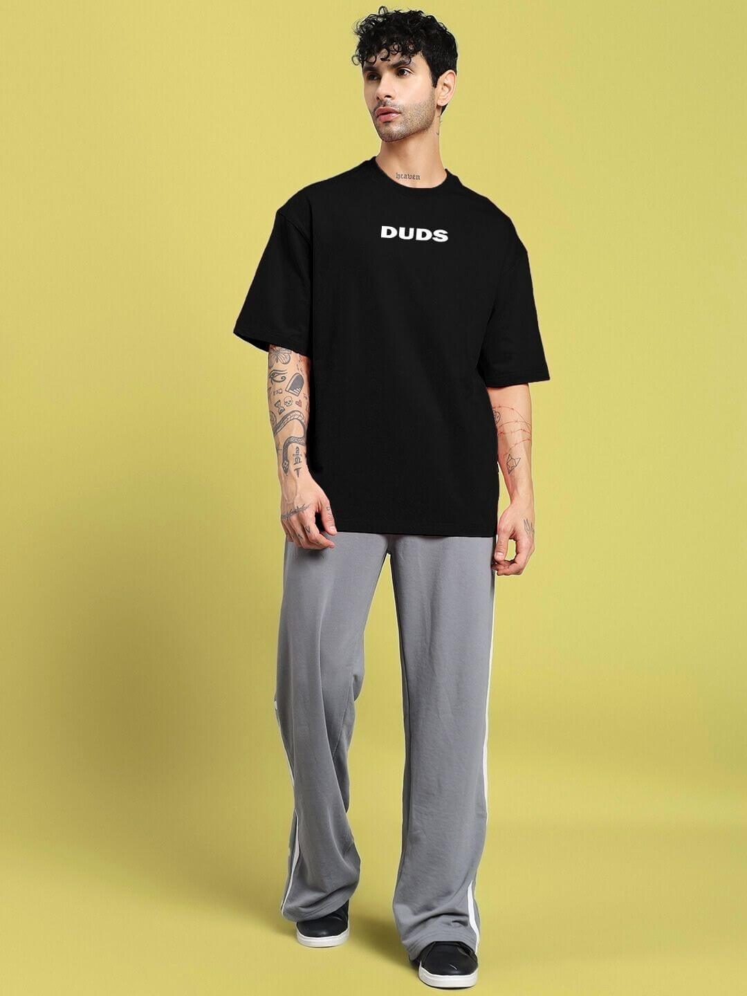 Man wearing DREAMLESS OVER-SIZED T-SHIRT BLACK, showcasing minimalist streetwear style, paired with gray pants against a yellow background.