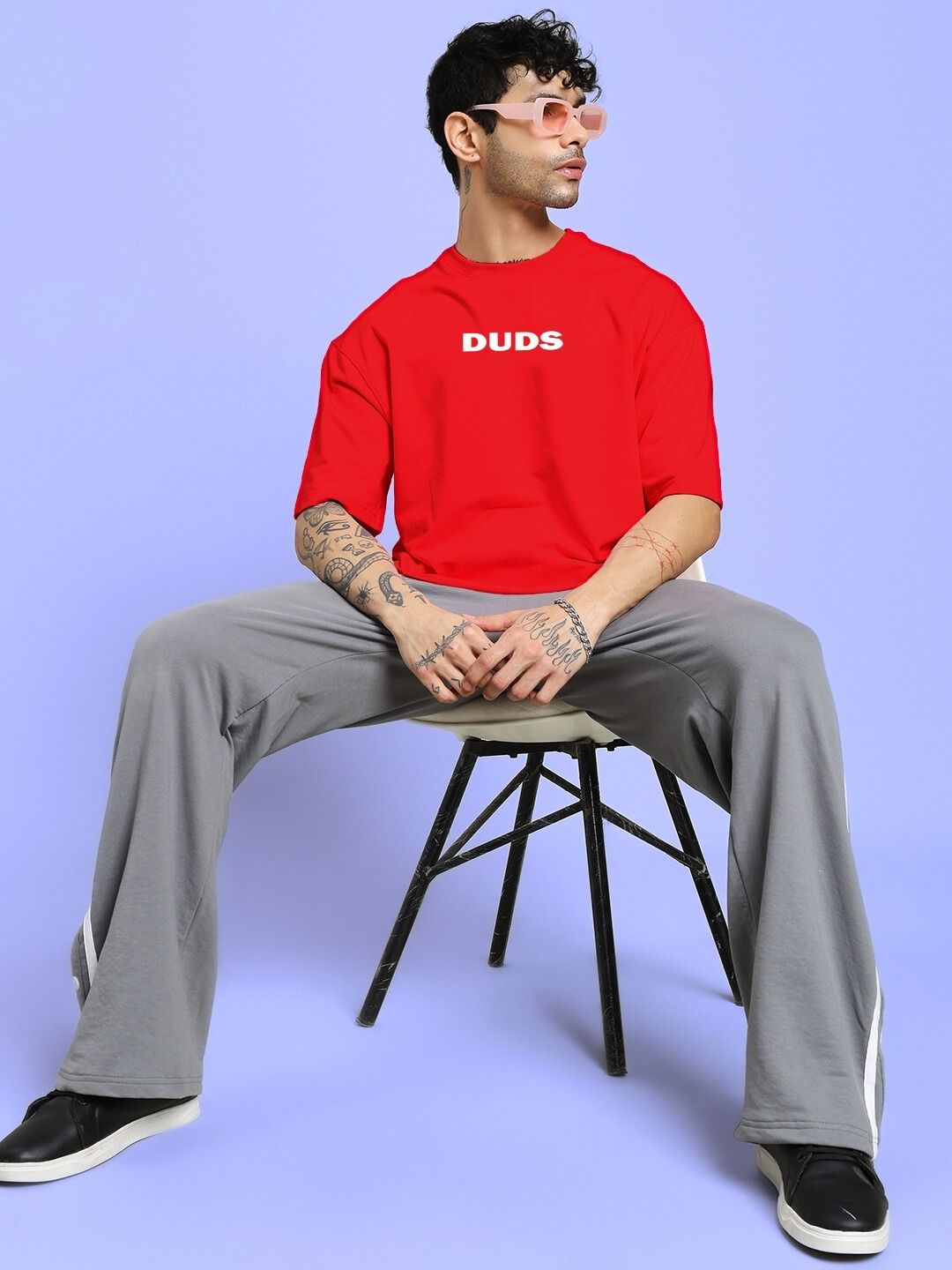 Model wearing DREAMLESS OVER-SIZED T-SHIRT RED with bold print, sitting on a stool, showcasing casual style.