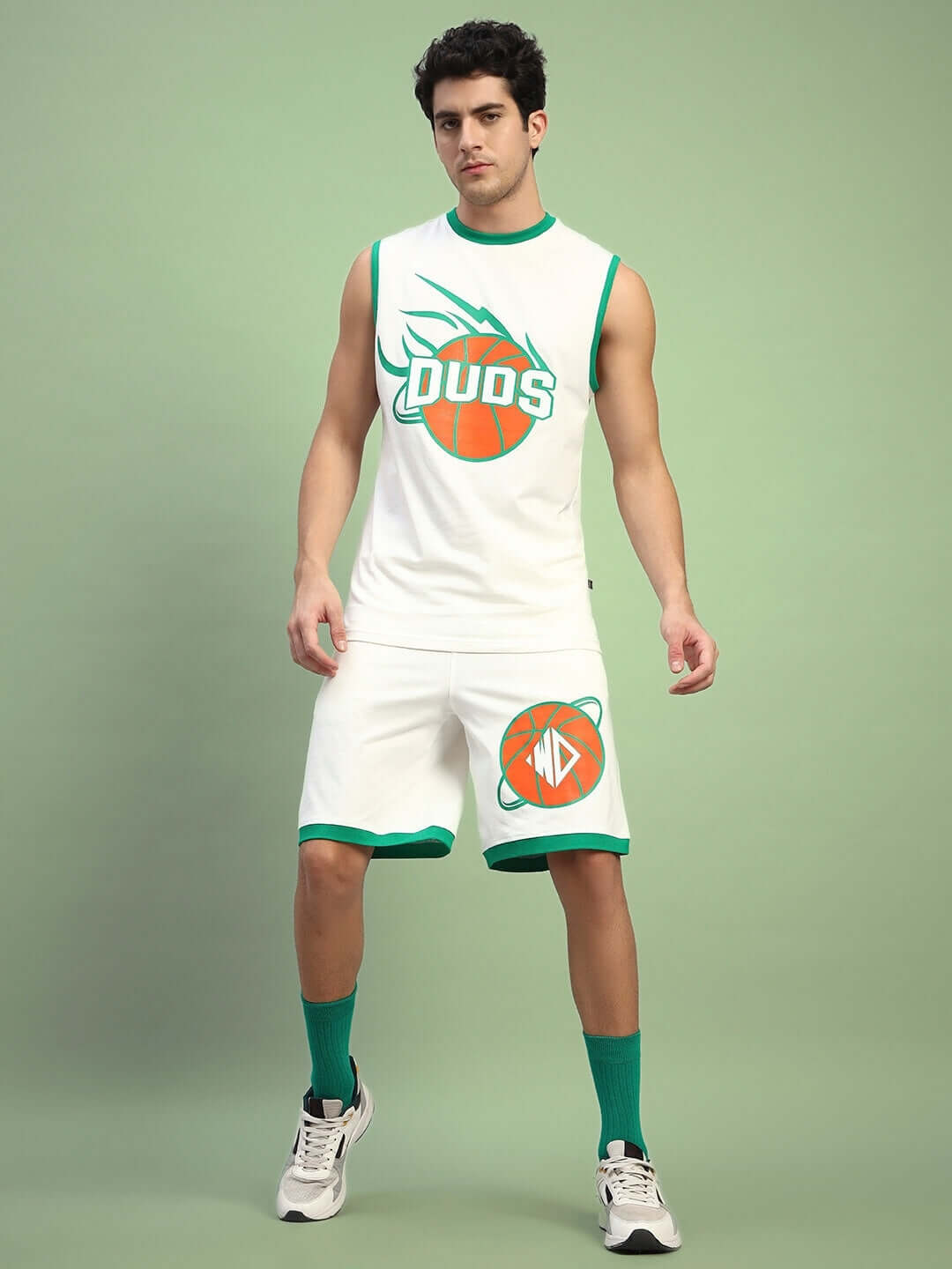 Model wearing DRIBBLE CO-ORD SET SLEEVELESS OFF WHITE, featuring sleeveless top and shorts with sporty design on a green background.