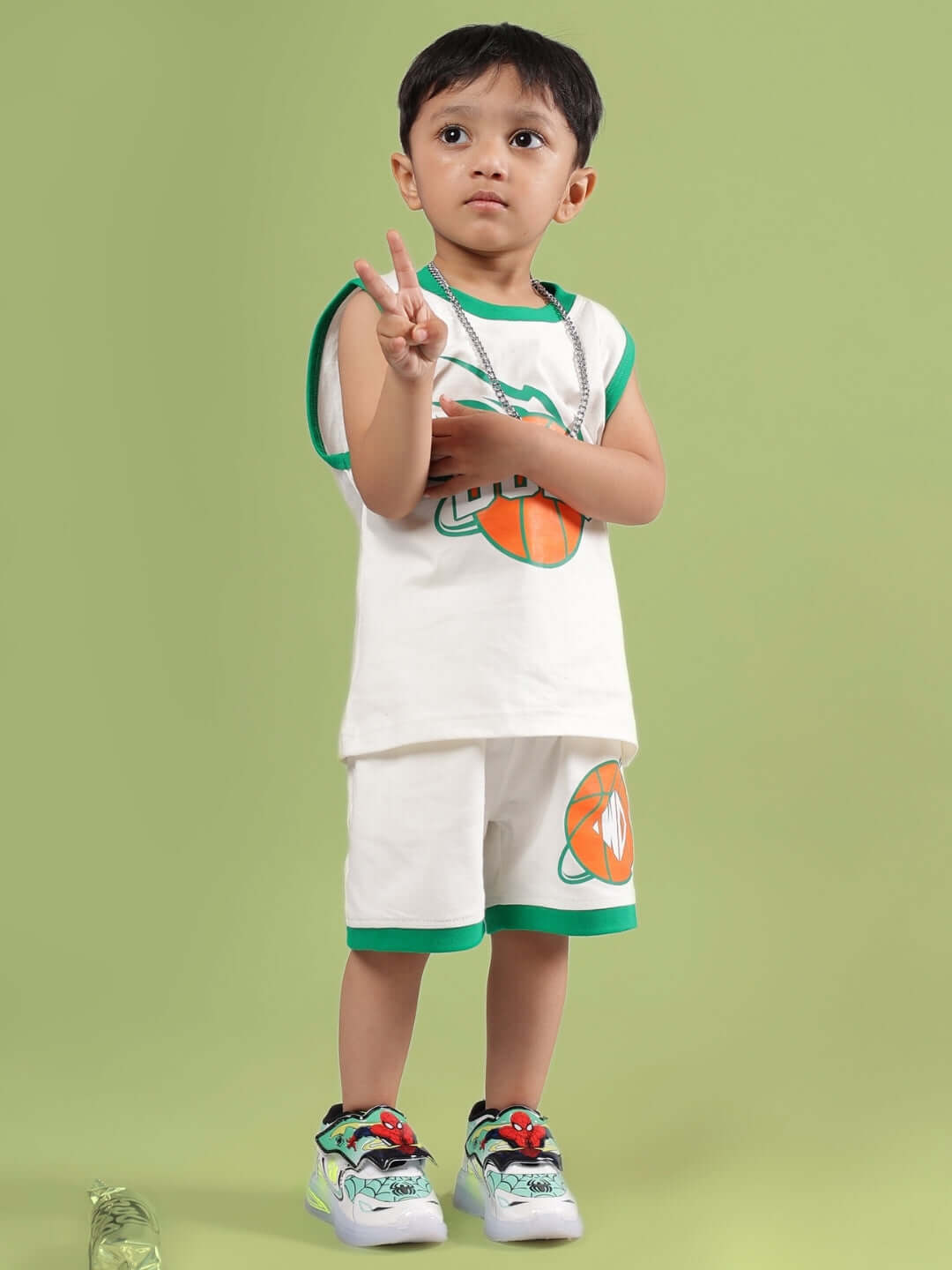 Child wearing DRIBBLE SHORTS FOR BOYS & GIRLS OFF WHITE with matching top, showing peace sign on a green background.