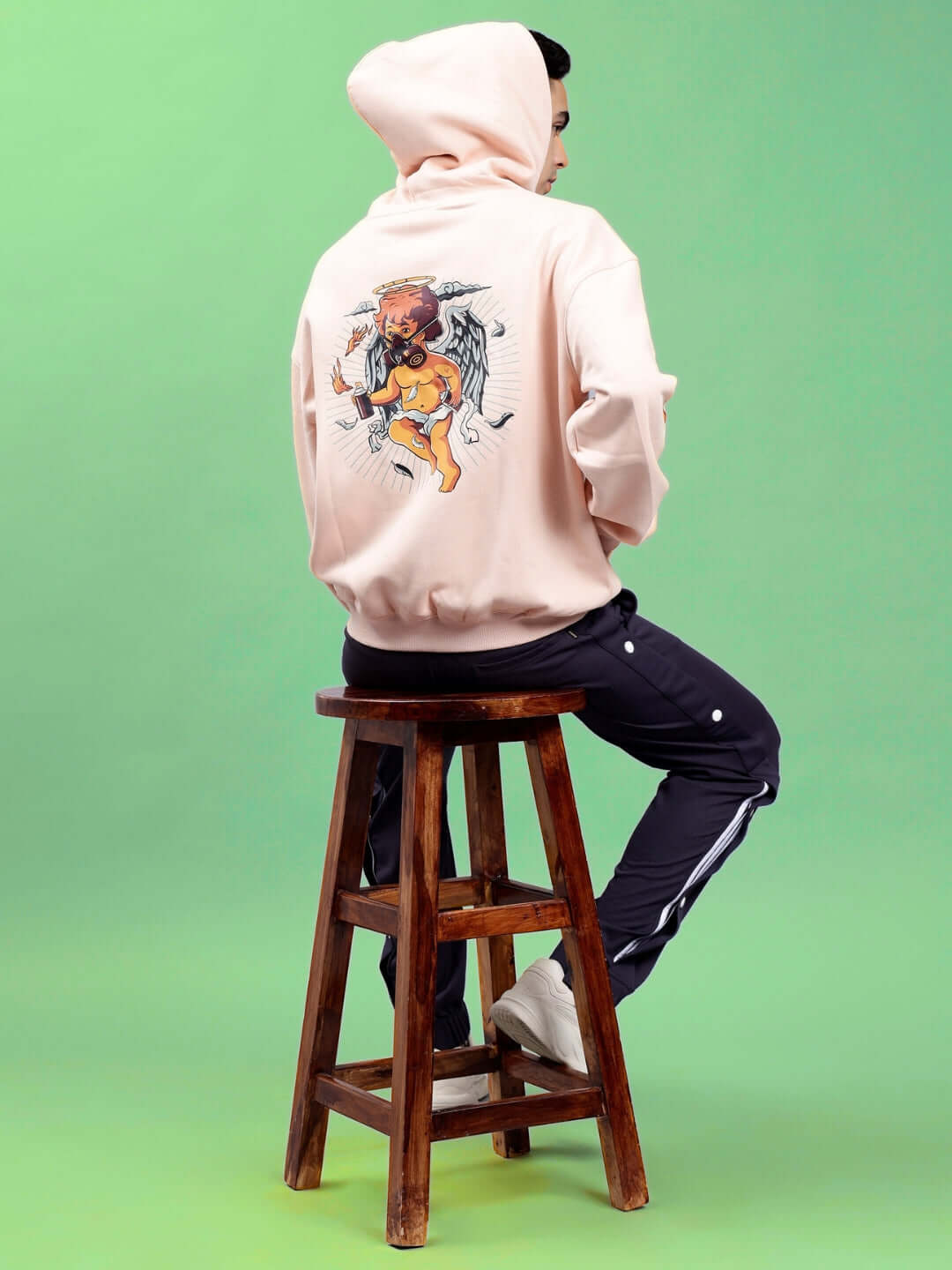 Person wearing DRIP CUPID OVERSIZED HOODIES BEIGE with graphic design, sitting on a stool, against a green background.