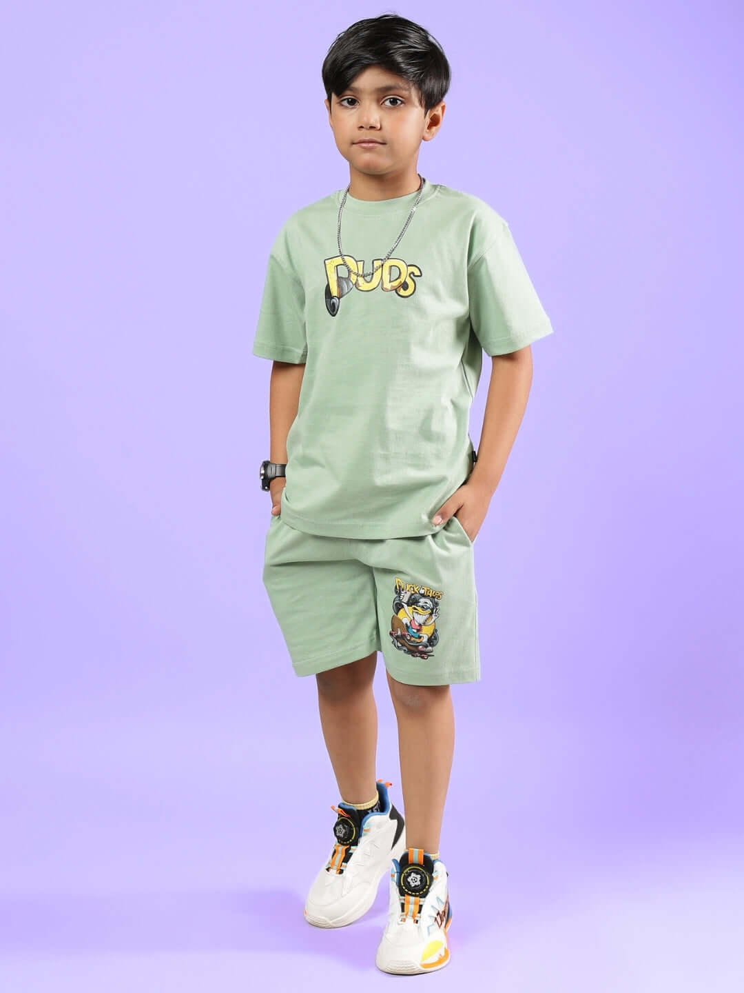 Child wearing DUCK TALES CO-ORD FOR BOYS & GIRLS NYANZA GREEN outfit with cartoon graphic, standing against a purple background.