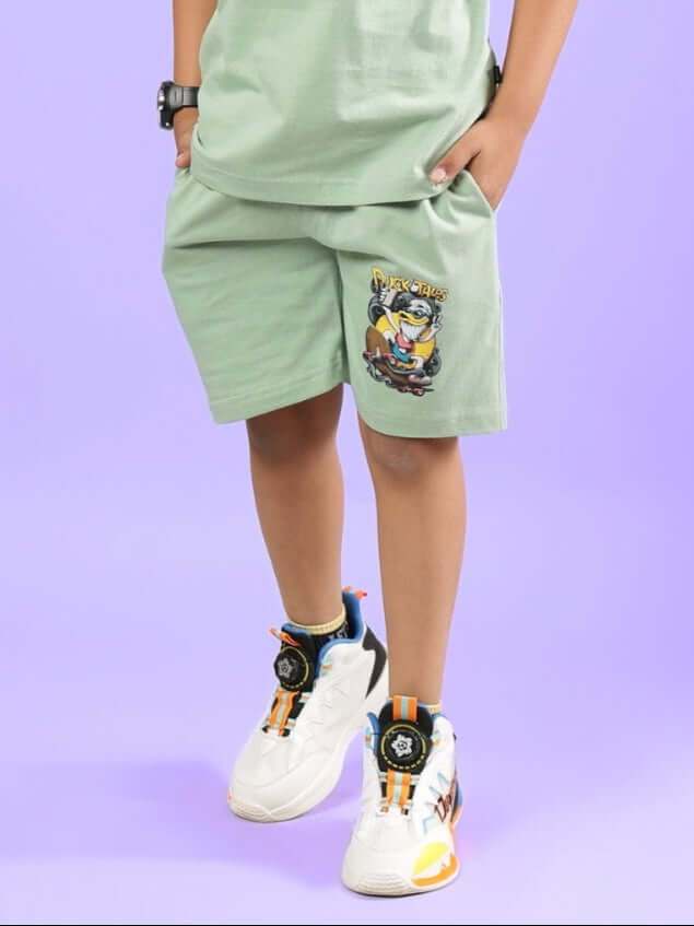 Child wearing DUCK TALES SHORTS FOR BOYS & GIRLS NYANZA GREEN with sneakers against a purple background.
