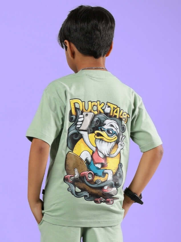 Boy wearing DUCK TALES T-SHIRT FOR BOYS GIRLS NYANZA GREEN with colorful graphic on back, in front of a purple background.