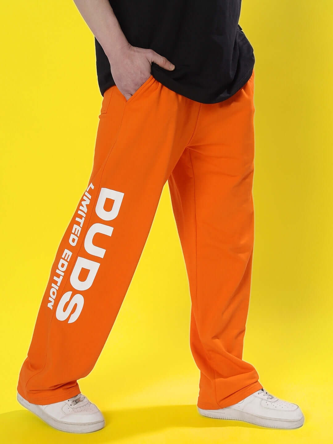 Model wearing DUDS Limited Edition Orange Joggers with bold text, showcasing vibrant style on a yellow background.