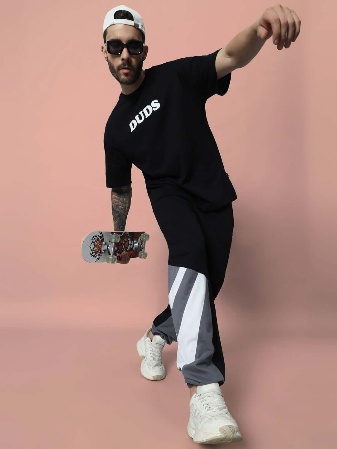 Model wearing DUDS relaxed fit joggers in black, holding skateboard, in dynamic pose on pink background.
