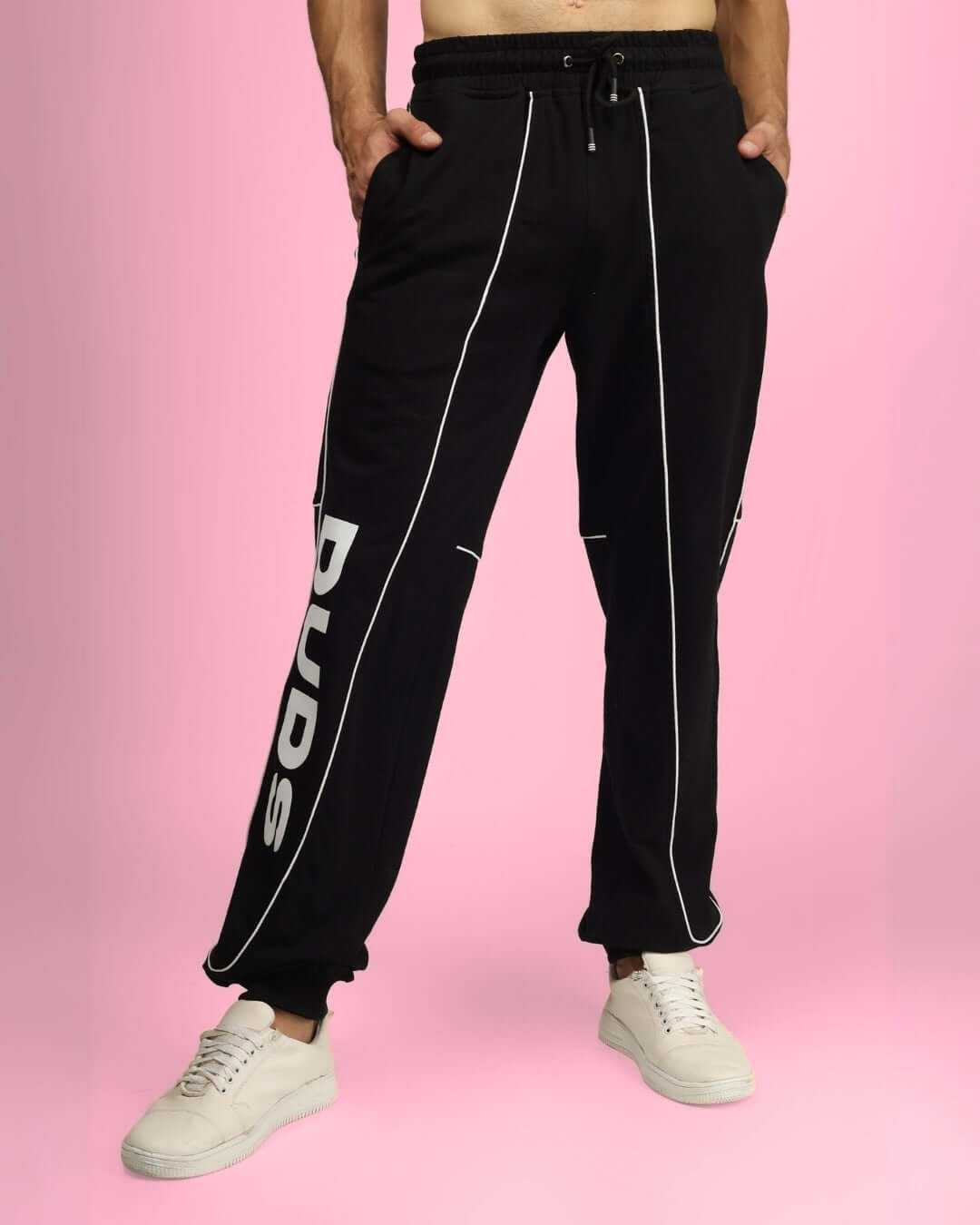 Man wearing DUDS Essential Joggers in black with white sneakers on a pink background.
