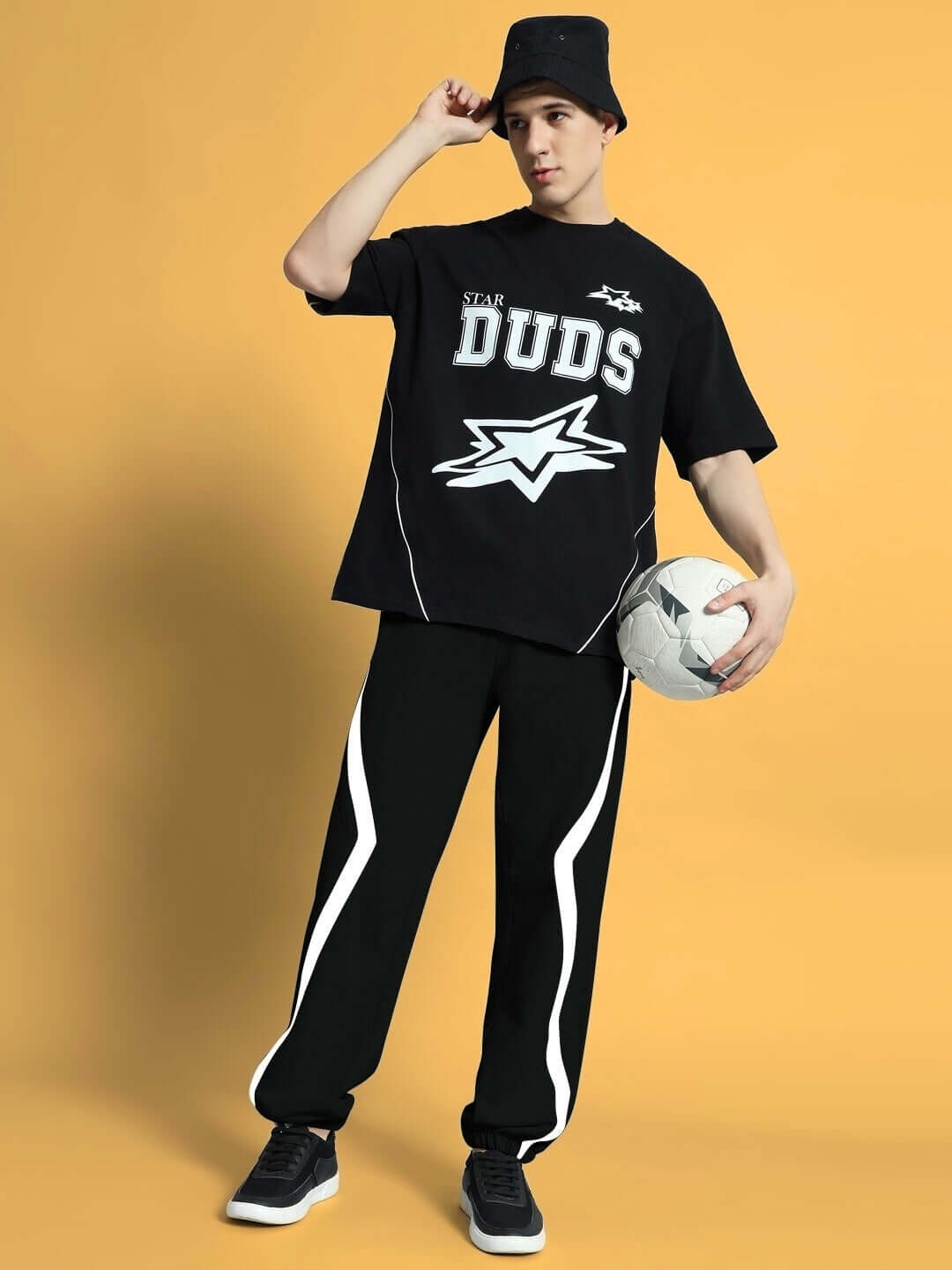 Man wearing DUDS FRAME JOGGERS in black with black t-shirt, holding a soccer ball, standing on orange background, casual athleisure style.
