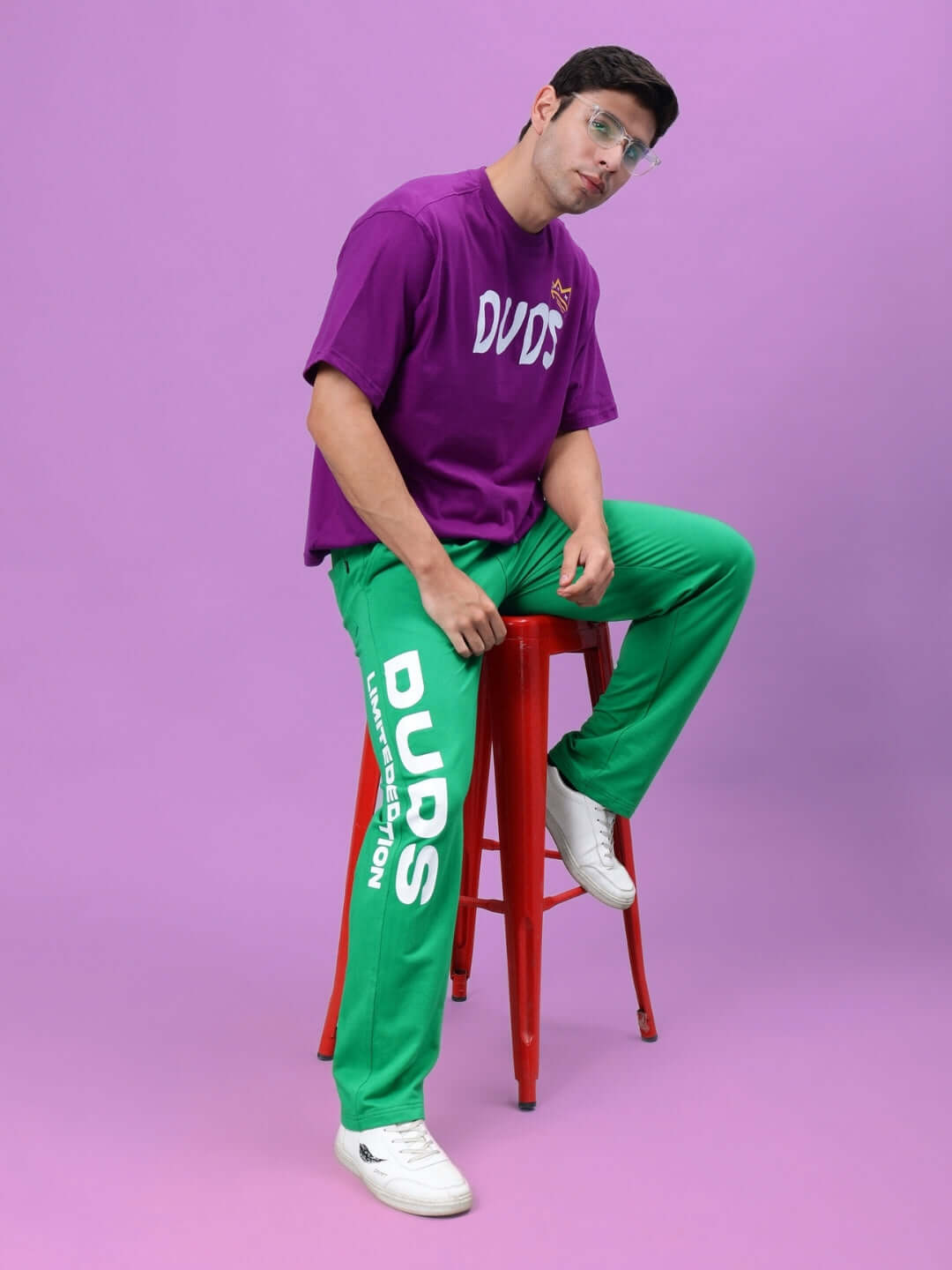 Man wearing DUDS Limited Edition Green Jogger and purple DUDS shirt sitting on red stool, showcasing trendy fashion style.