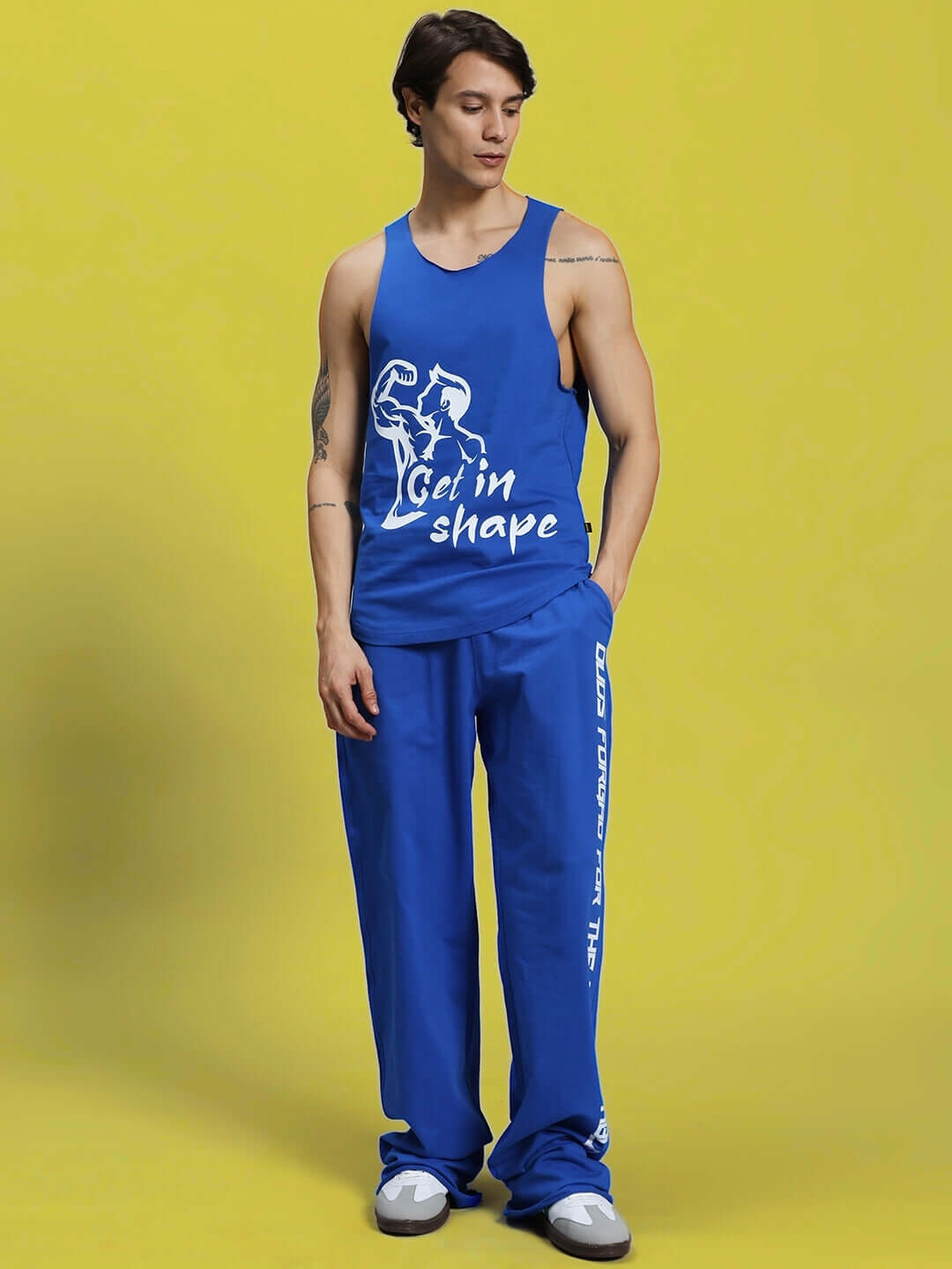 Man wearing DUDS Swift Jogger in royal blue with "Get in Shape" tank top, standing against a yellow background.