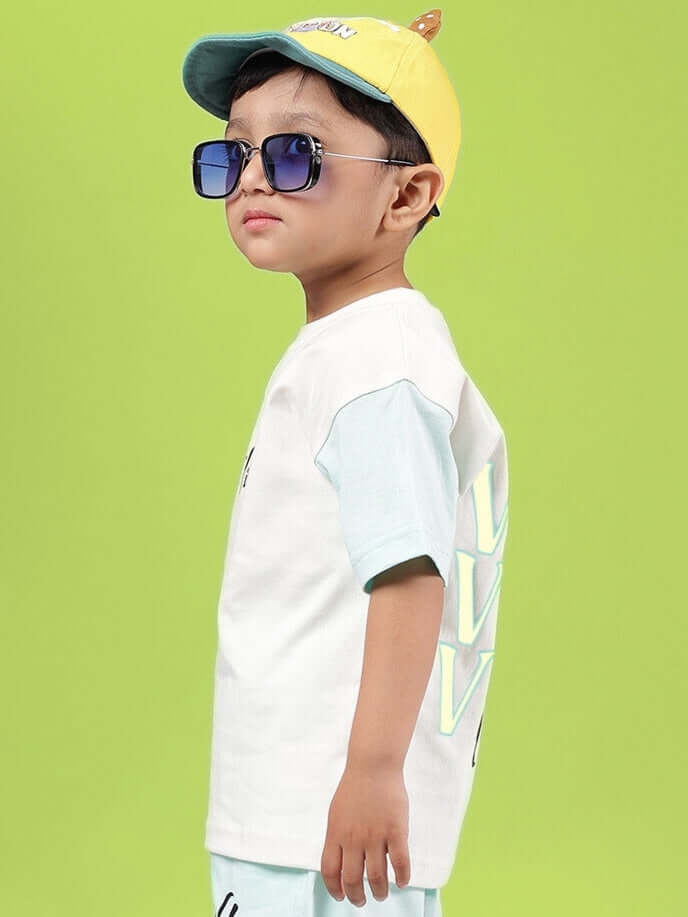 Young child wearing off white-blue DUDS Vibes T-shirt, yellow cap, and sunglasses, standing against a bright green background.