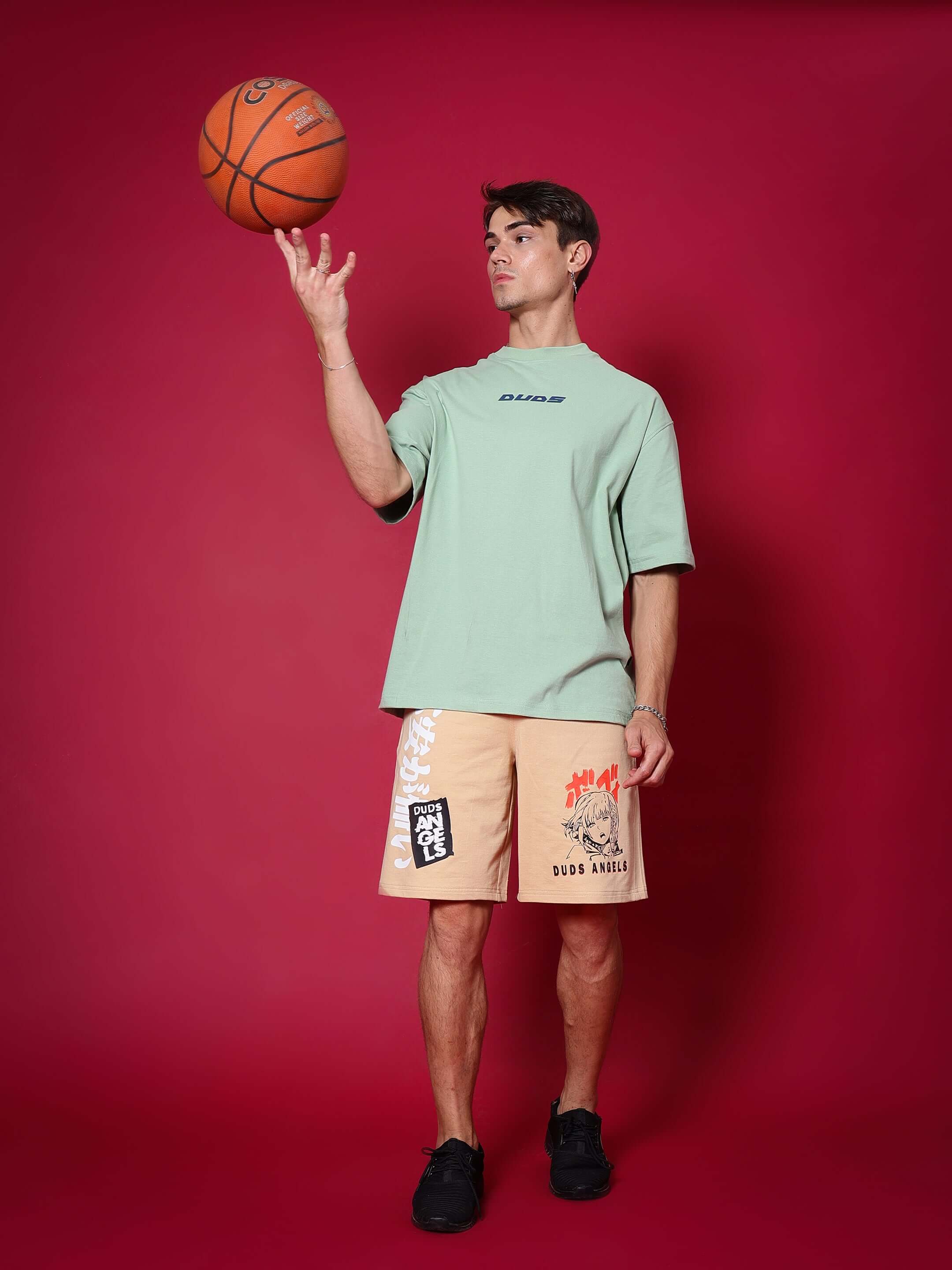 Man wearing DUDS Anime Girl Regular Fit Shorts in light orange with graphic prints, standing on a red background while tossing a basketball.
