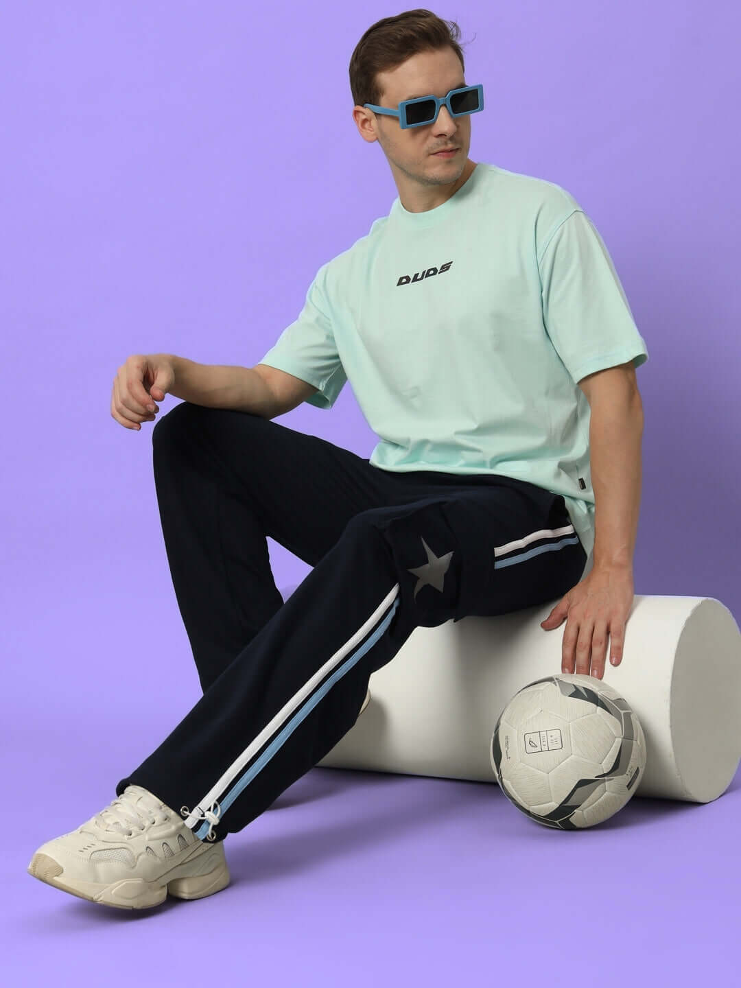 Model wearing DUDS Force Joggers in Navy Blue, sitting stylishly with a soccer ball. Tailored fit, available in custom sizes.