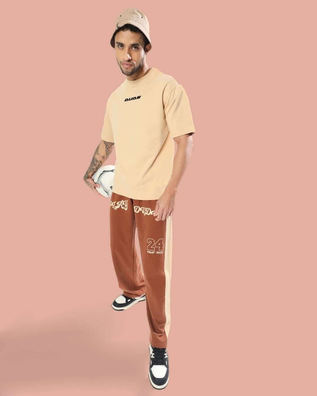 Man wearing DUDS PLAY baggy joggers in brown-beige, paired with a tan shirt and bucket hat, standing against a peach background.
