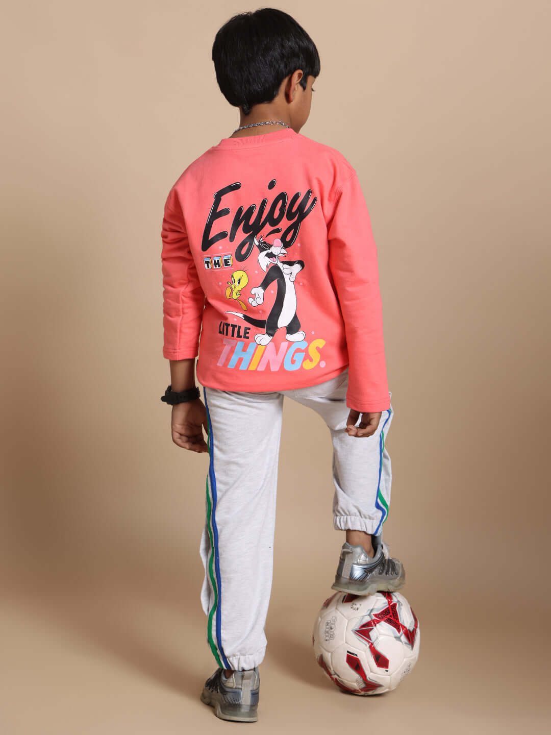 Boy wearing mélange grey DUDS PRISM colorful stripe jogger and red graphic top, standing with a soccer ball on beige background.