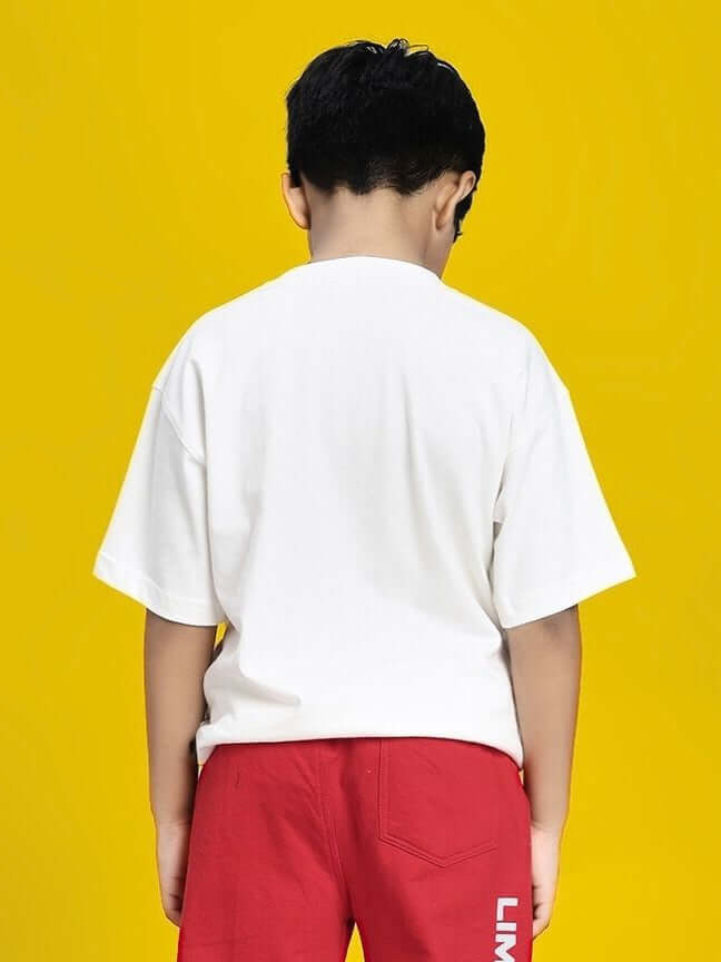 Child wearing a DUMBO T-SHIRT FOR BOYS AND GIRLS WHITE with red shorts, standing against a yellow background, back view