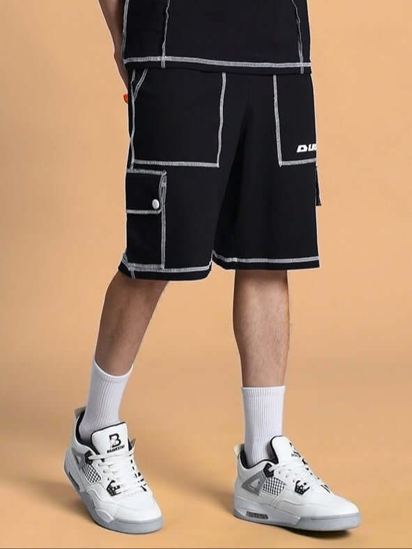 Model wearing DYNAMIC REGULAR FIT SHORTS BLACK with white sneakers on beige background.