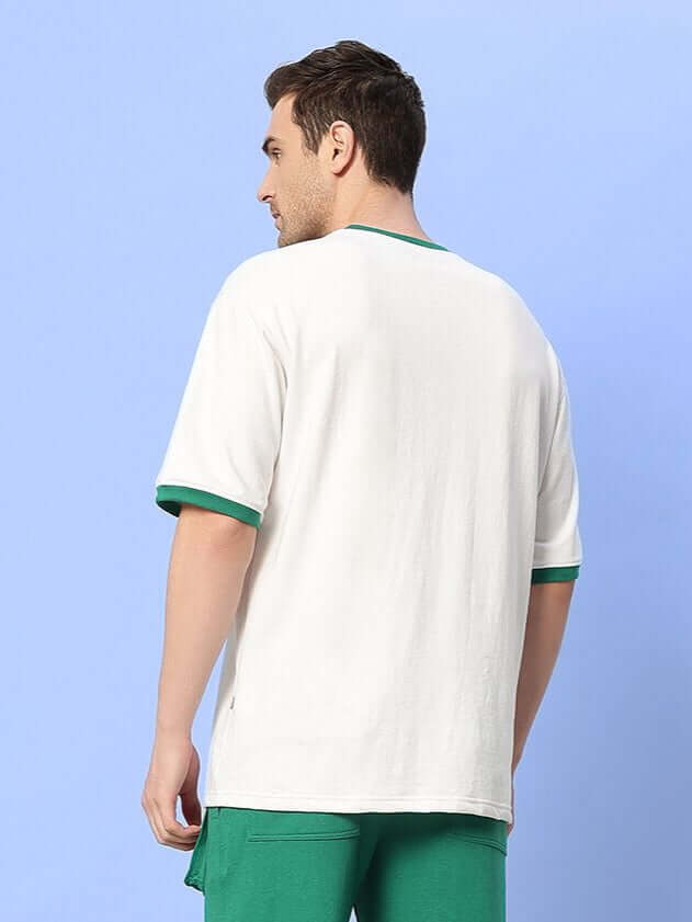 Man wearing EASTON BOXY FIT T-SHIRT WHITE-GREEN, showcasing back view of the white tee with green trim against blue background.