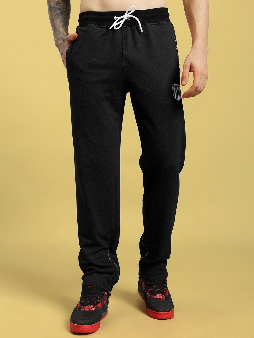 Man wearing ELEGANT RELAXED-FIT JOGGERS BLACK with red-accented sneakers on a beige background.