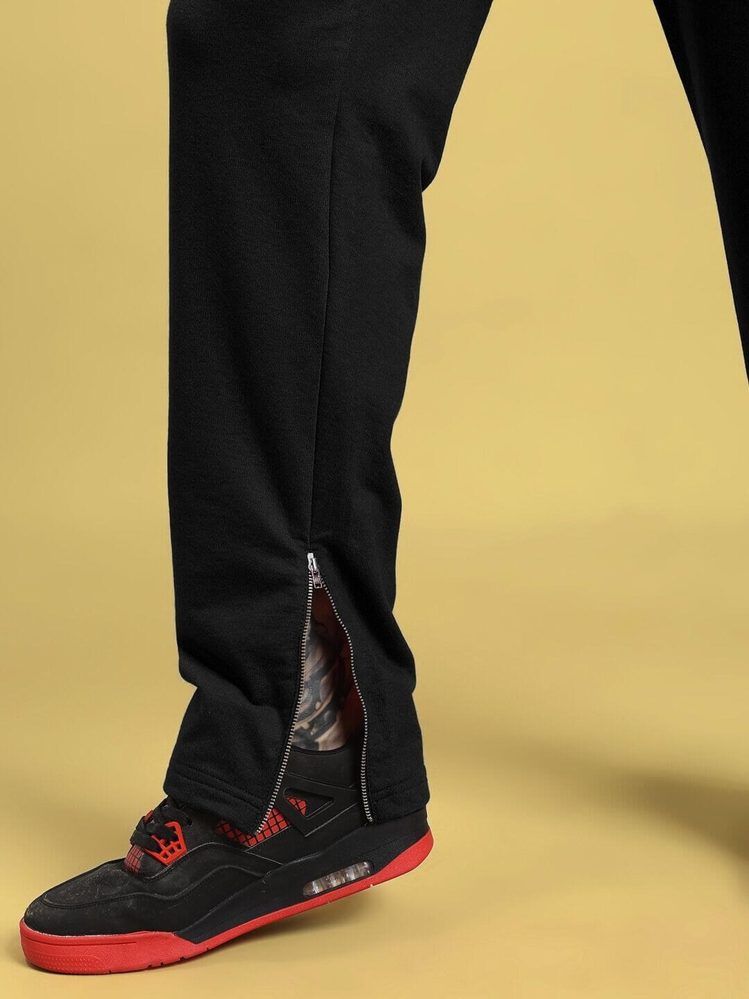 Close-up of legs wearing ELEGANT RELAXED-FIT JOGGERS BLACK with sneakers, showcasing sleek silhouette and zipper detail, on yellow background.