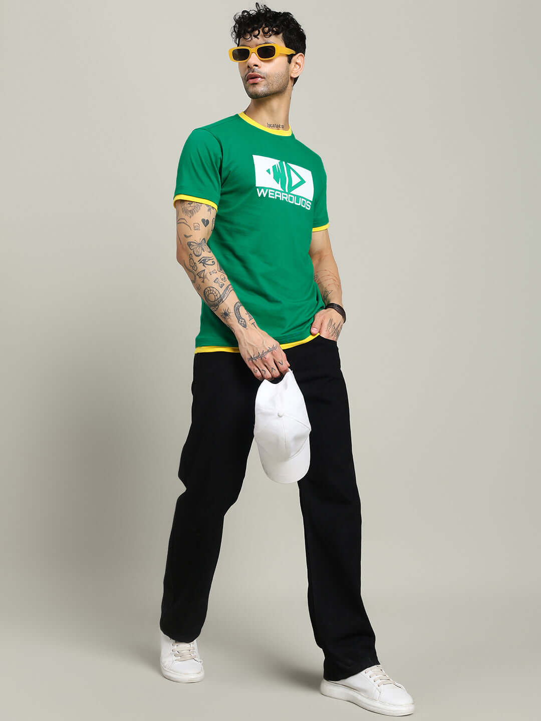 Man wearing ELMER REGULAR FIT T-SHIRT GREEN paired with black pants and yellow sunglasses, holding a white cap.