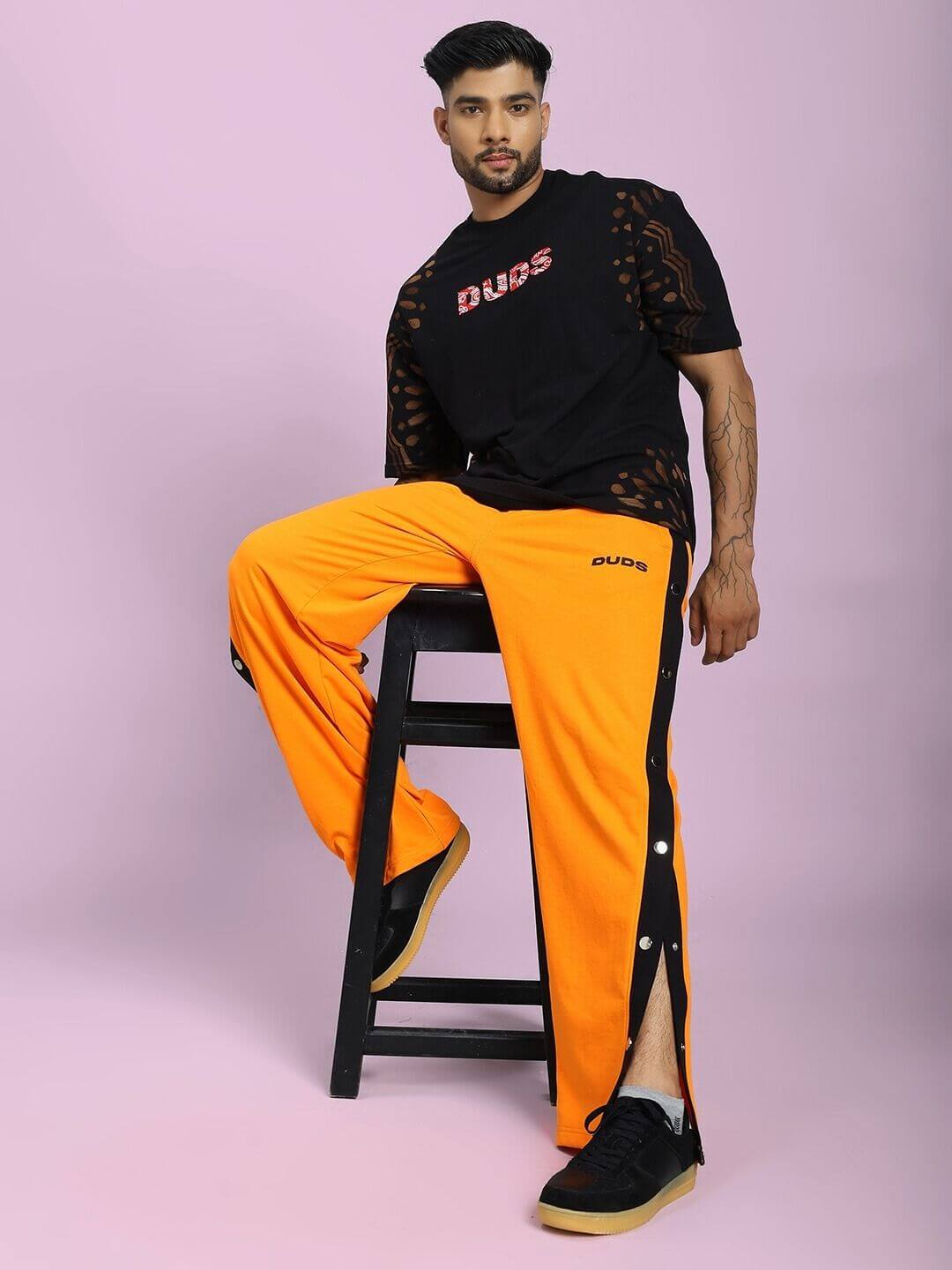 Person wearing EMBER BOLT SIDE JOGGER IN ORANGE with side stripe, seated on stool, showcasing vibrant style and comfortable fit.