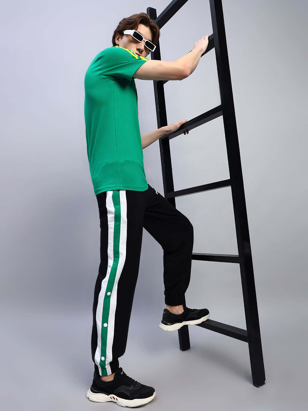 Man wearing EMIRATI STRIPES JOGGERS BLACK-GREEN with green stripes, standing near a ladder, showcasing trendy streetwear fashion.