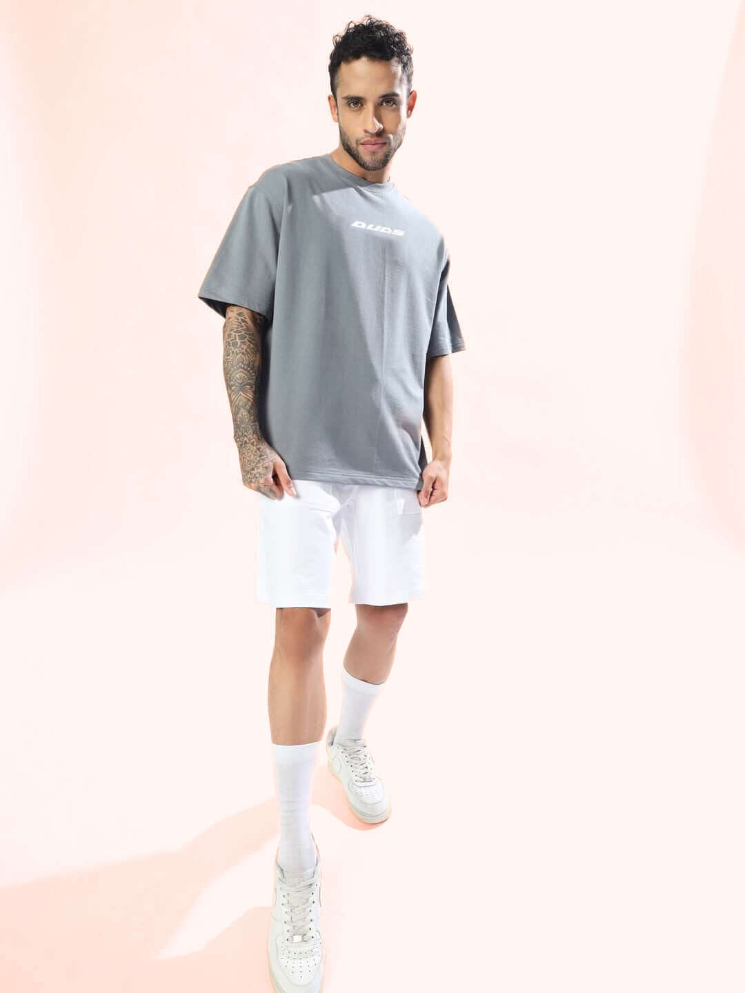 Model wearing an oversized grey t-shirt with white shorts and white sneakers, showcasing street style fashion on a pastel background.
