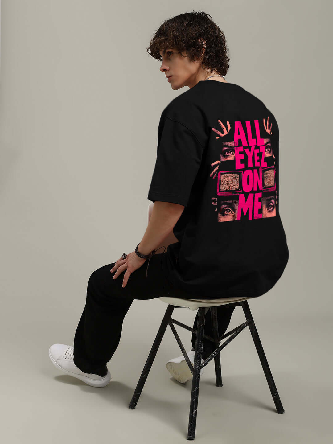 Model wearing EYECONIC OVERSIZED PRINTED T-SHIRT BLACK with vibrant graphic design, seated on chair, showcasing bold street-style fashion.