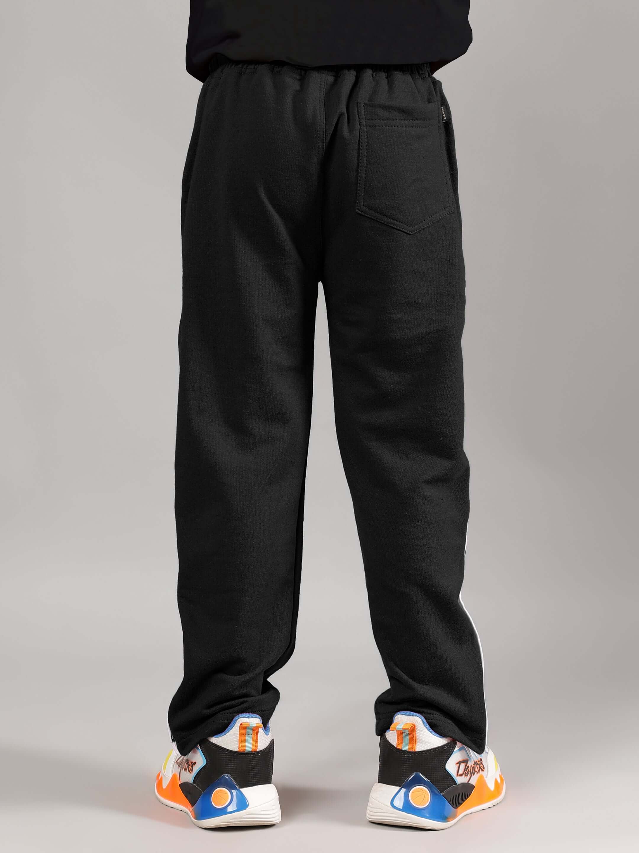 Child wearing EZEL JOGGER FOR BOYS & GIRLS BLACK with colorful sneakers, back view, showing elastic waistband and pockets. Comfortable fit.