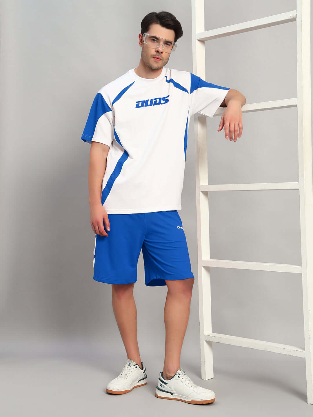 Man wearing EZEL REGULAR FIT COTTON SHORTS BLUE with a white and blue sports jersey, leaning against a white ladder.