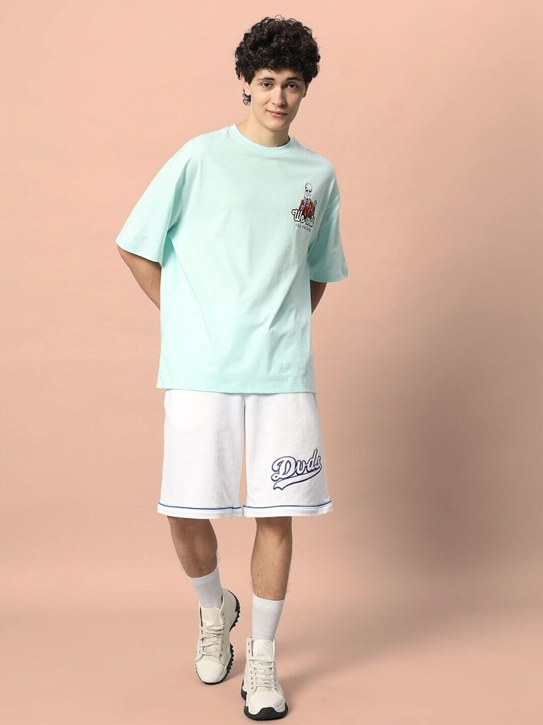 Man wearing CALL YOU BACK OVER-SIZED T-SHIRT ICE BLUE and white shorts, standing against a peach background.