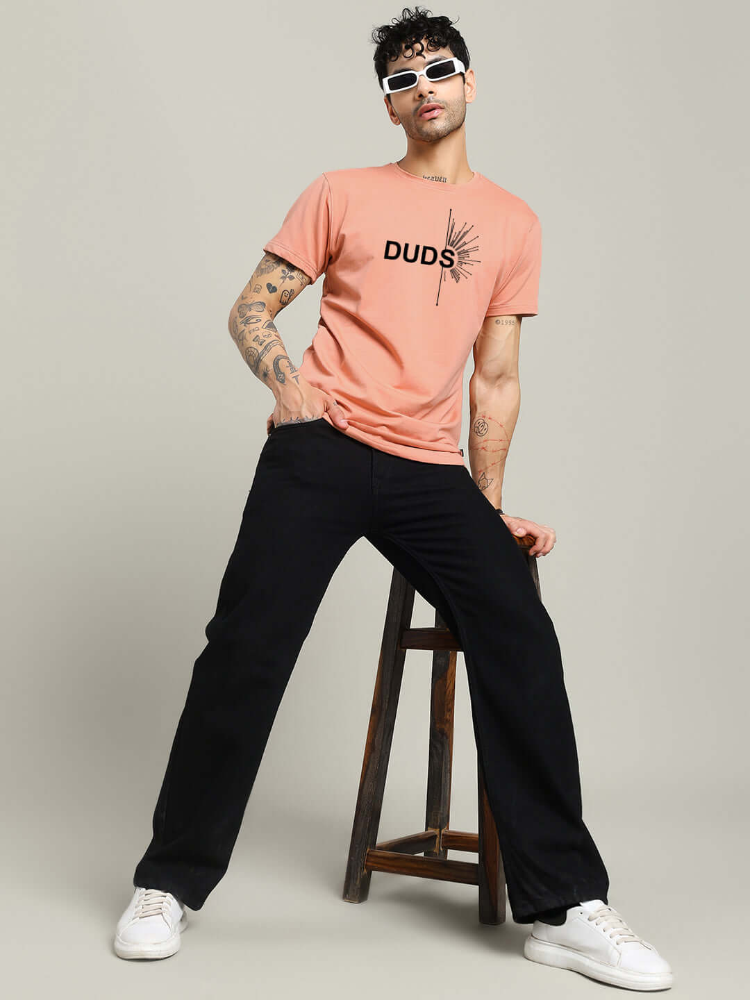 Man wearing peach Fallen Angle T-shirt, regular fit, 100% cotton, seated on wooden stool, styled with sunglasses and black pants.