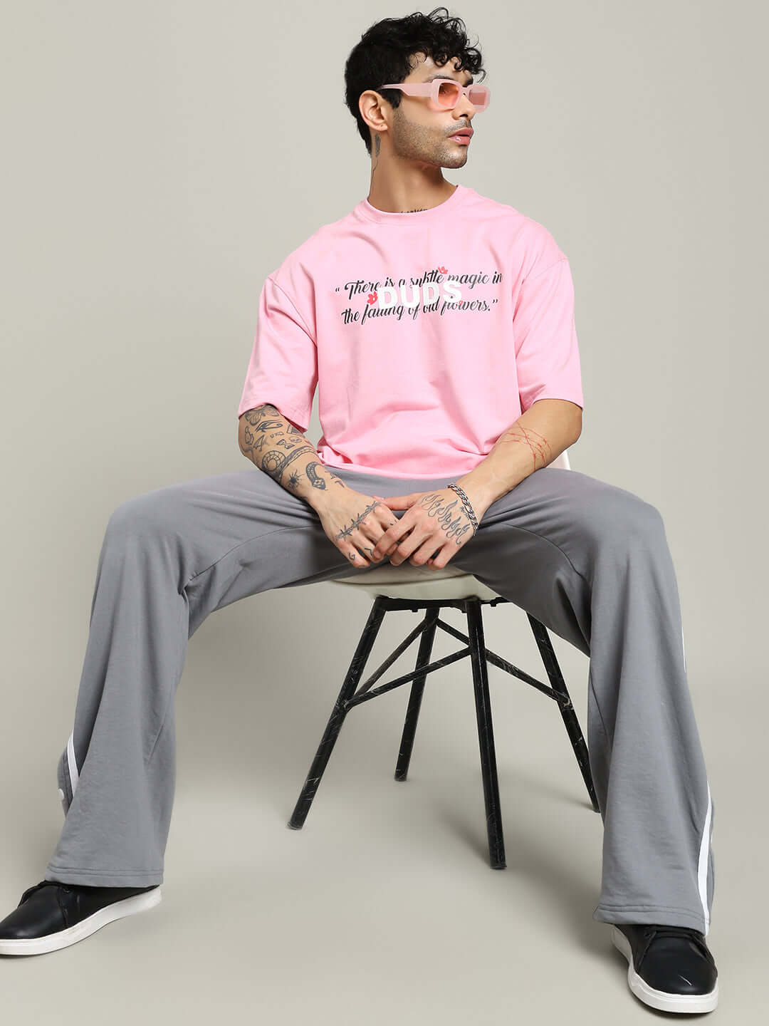 Man wearing FALLING FLOWER T-SHIRT PINK sitting on a chair with casual gray pants and sneakers.
