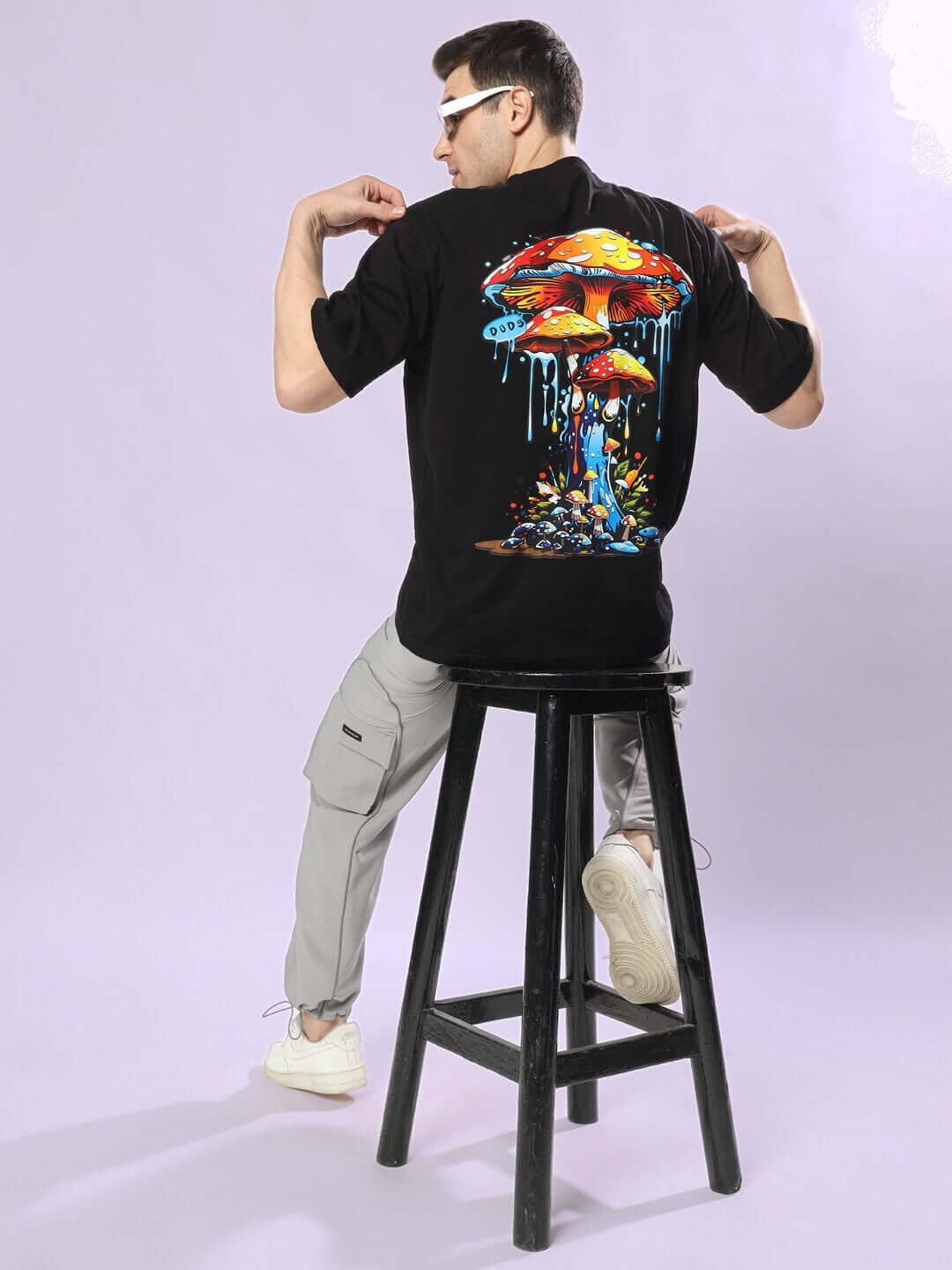 Person wearing a Fantasy Mushroom Over-sized T-shirt in black, sitting on a stool, showcasing vibrant mushroom graphic on the back.