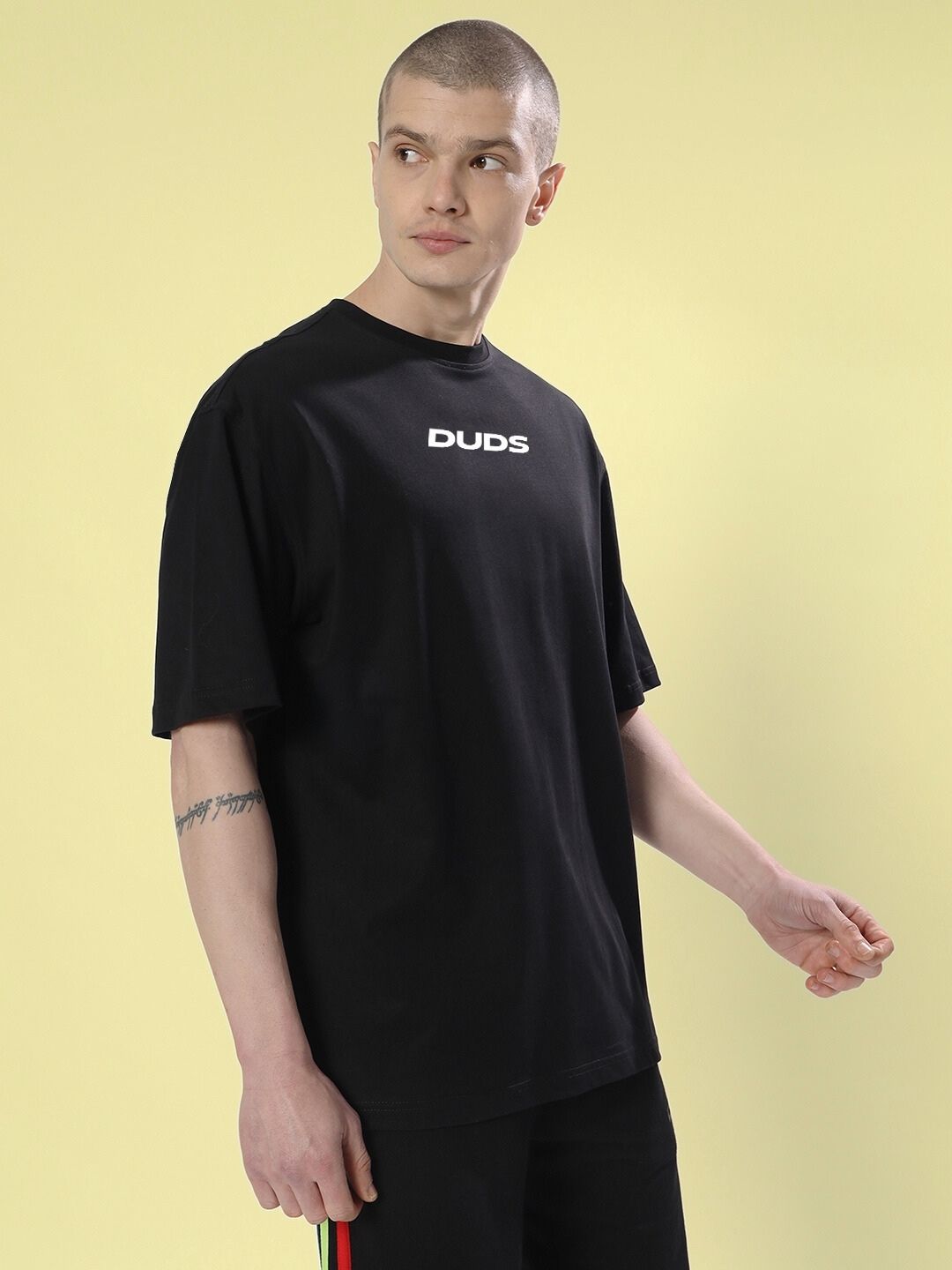 Man wearing FARARI PRINTED OVERSIZED T-SHIRT BLACK with logo, standing against yellow background showcasing casual style.