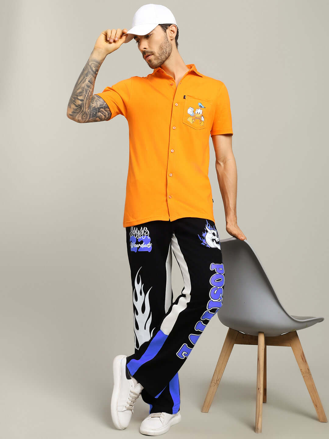 Model in orange shirt and black-blue joggers with graphic designs, white sneakers, and cap, posing with chair in studio setting.
