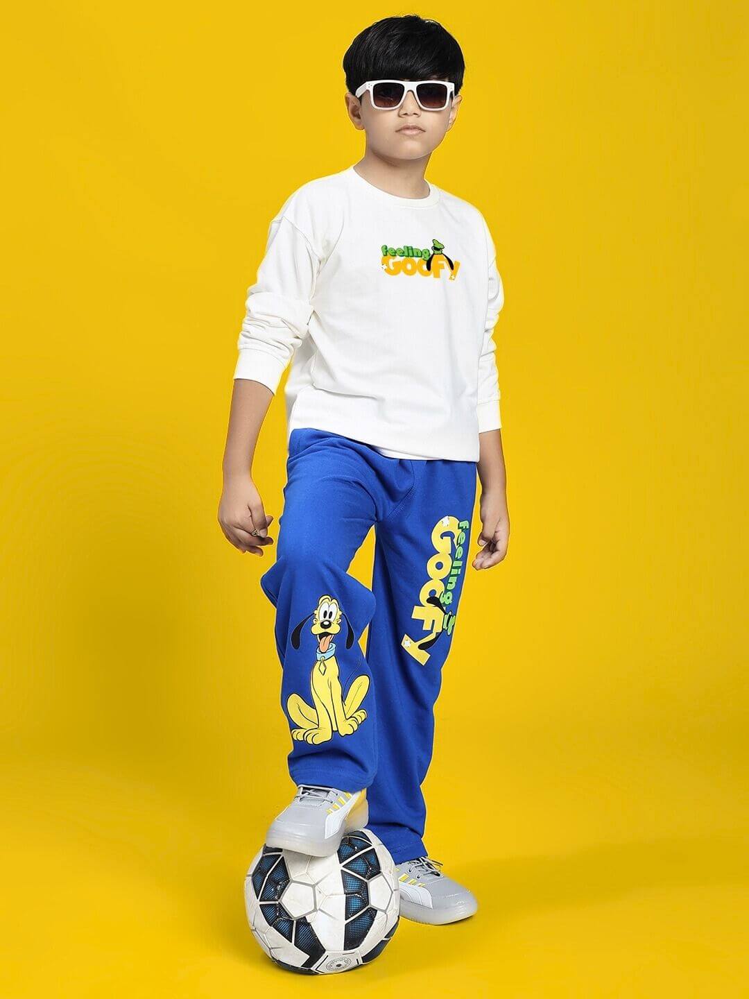Child wearing FEELING GOOFY JOGGER FOR BOYS AND GIRLS BLUE with a soccer ball and white sneakers, on a yellow background.