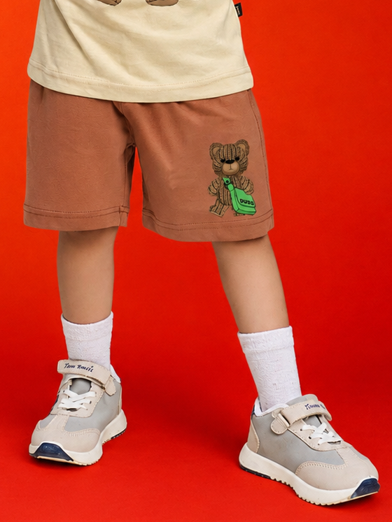 LITTLE EXPLORER TEDDY SHORTS FOR BOYS GIRLS BROWN worn by a child against a bright red background.