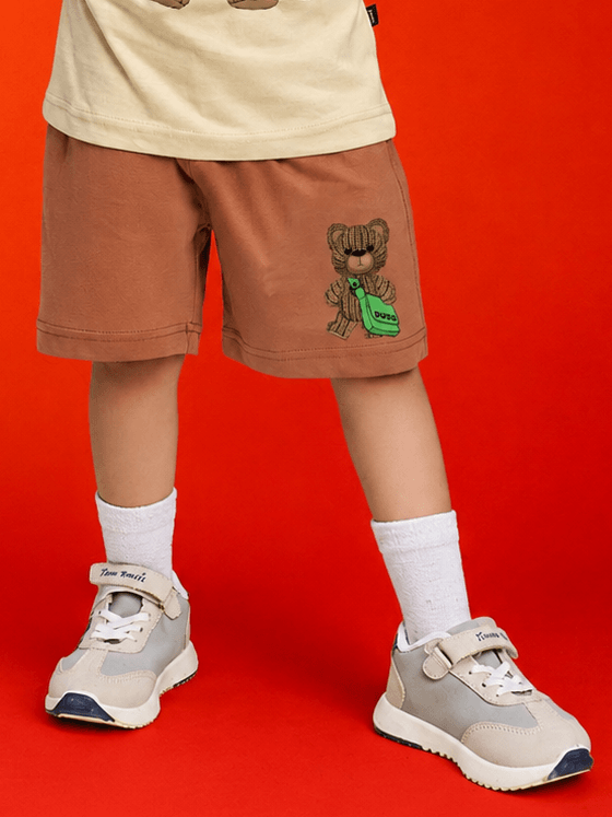 LITTLE EXPLORER TEDDY SHORTS FOR BOYS GIRLS BROWN worn by a child against a bright red background.