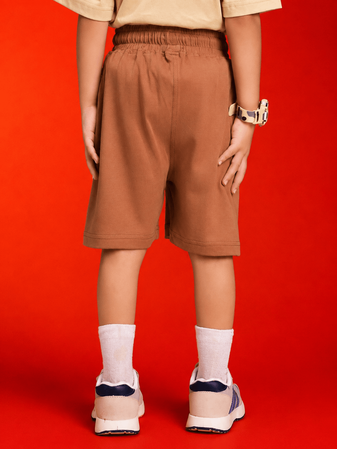 Little Explorer Teddy Shorts for Boys Girls Brown showcased on a child from the back against a bright red background.