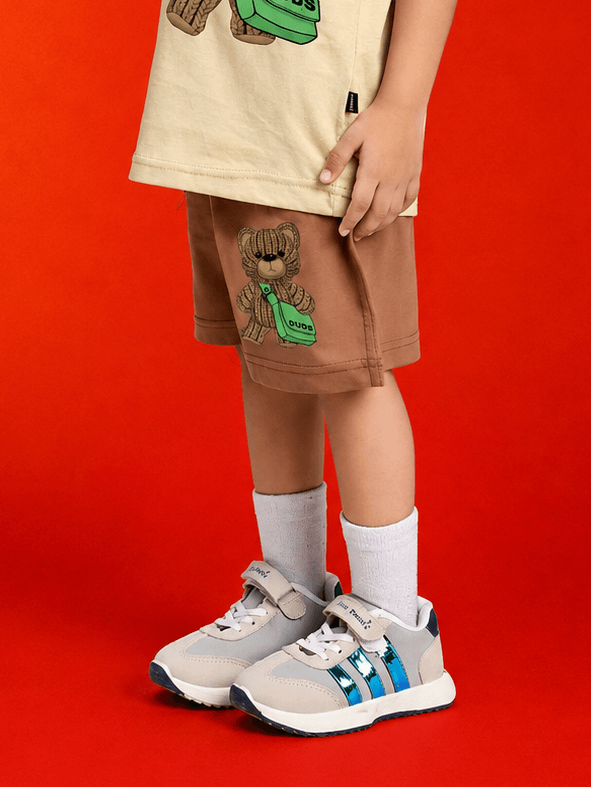 A child wearing LITTLE EXPLORER TEDDY SHORTS FOR BOYS GIRLS BROWN with cute teddy bear design and casual sneakers.