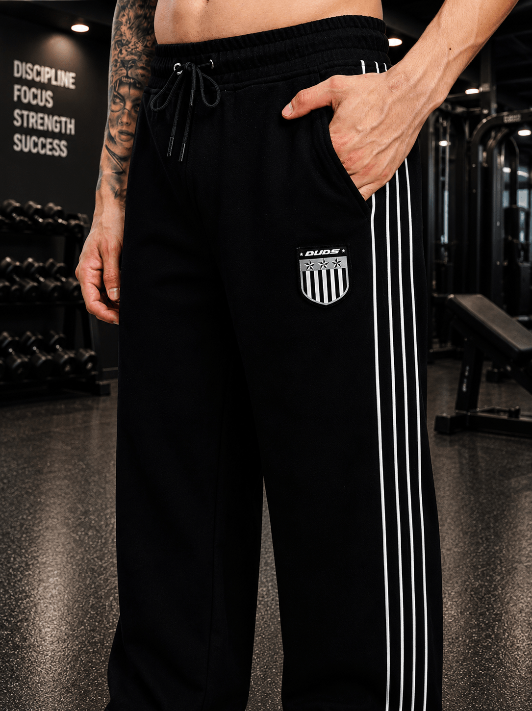 Model wearing BLACK OUT-FIT JOGGERS BLACK with stylish white stripes and logo in a gym setting.