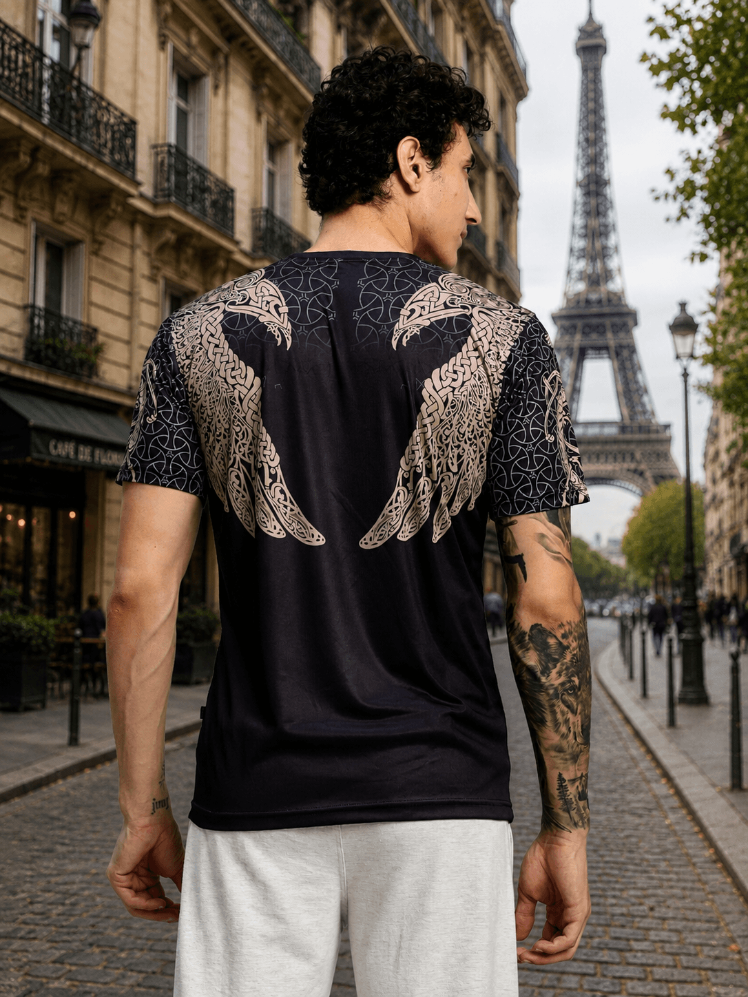 Back view of a man wearing AURA OF ASGARD REGULAR T-SHIRT BLACK with intricate wing design, Eiffel Tower in the background.