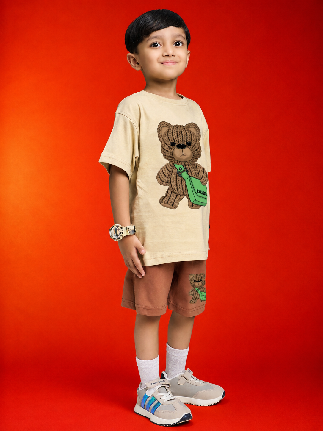 LITTLE EXPLORER TEDDY CO-ORD FOR BOYS GIRLS BEIGE BROWN modeled by a smiling boy against a vibrant red background.