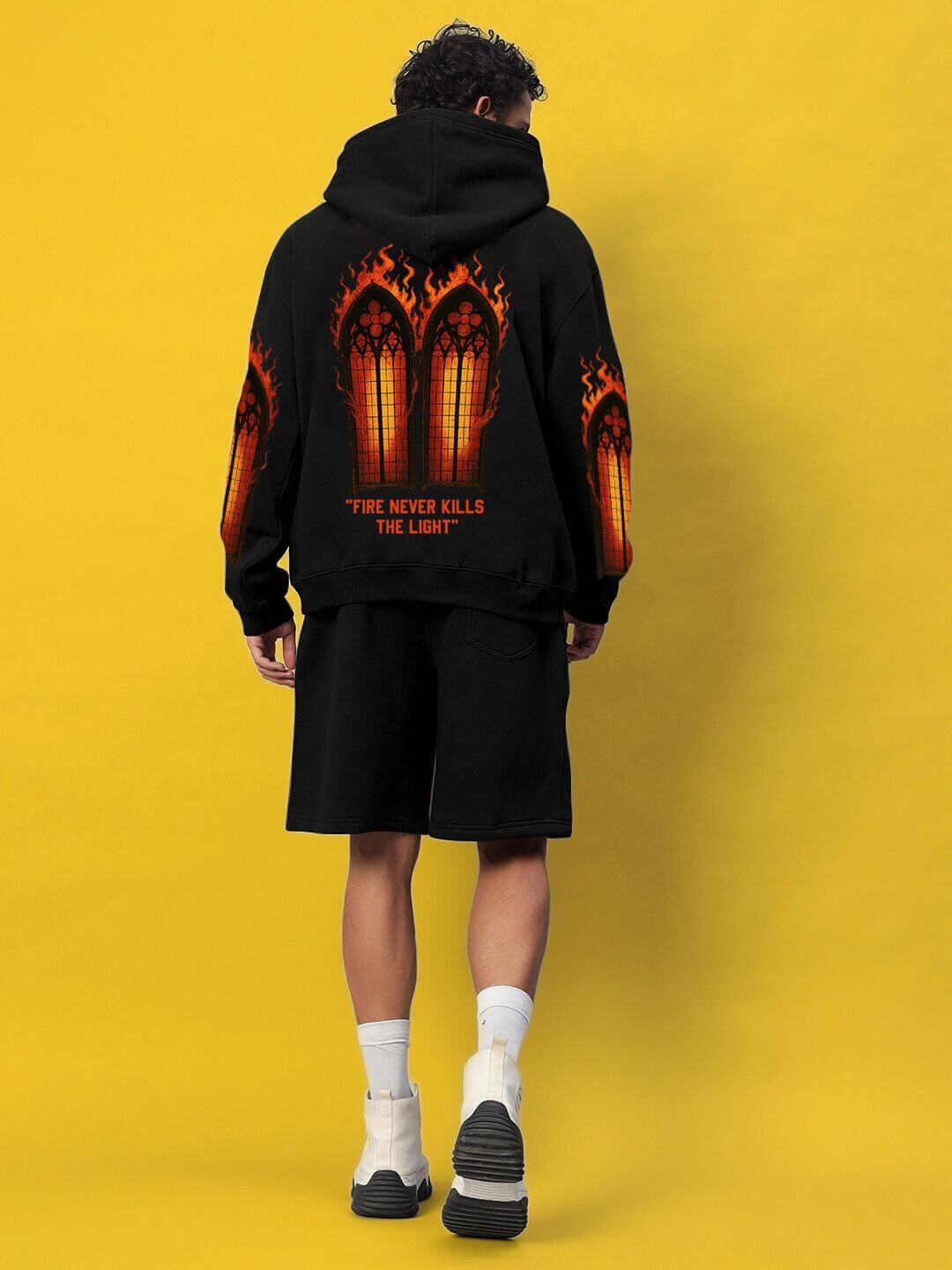 Man wearing FIRE NEVER KILLS THE LIGHT FLEECE CO-ORD BLACK, showing bold design with fiery graphics on back against yellow background.