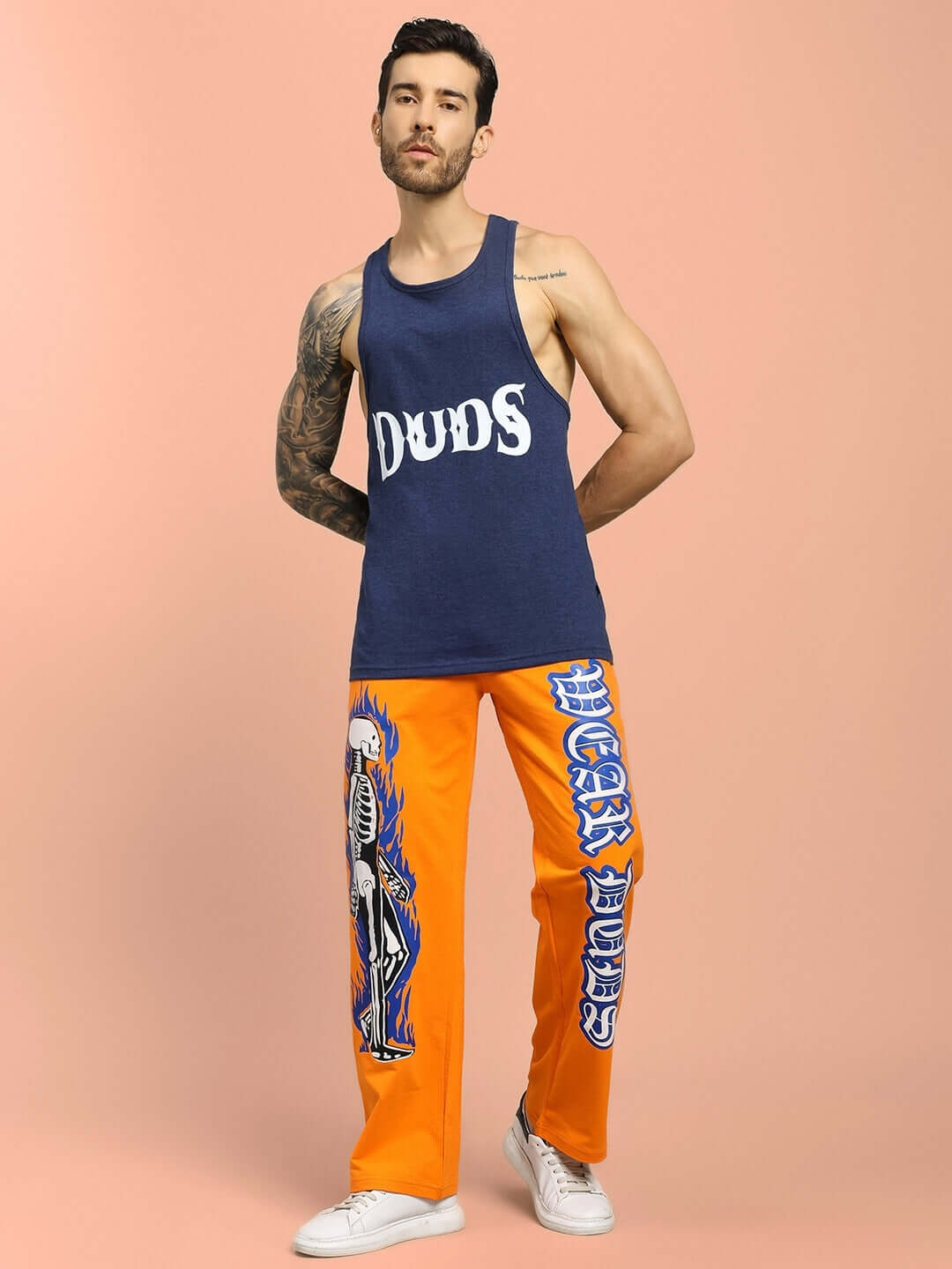 Man wearing FLAMING SKELETON PRINTED RELAXED FIT JOGGER ORANGE and navy tank top, posing against a peach background.